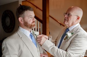 Older man helping groom adjust his tie before wedding ceremony, both wearing tan suits.