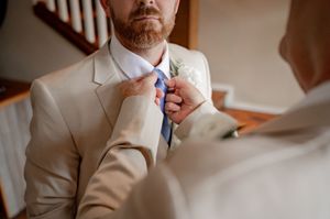 Groom in beige suit with blue tie and boutonniere being adjusted by someone during wedding preparations.