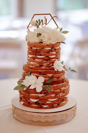 Three-tier naked wedding cake stacked with cookies, white flowers, greenery, and a gold hexagon Mr. & Mrs. topper.
