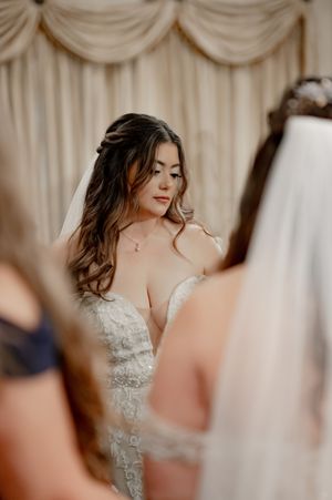 Beautiful bride in lace wedding gown with veil stands surrounded by bridesmaids before ceremony.