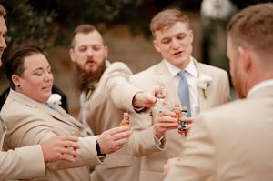 Wedding party in tan suits toasting with mini liquor bottles outdoors during celebration.