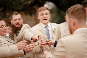 Groomsmen in beige suits raising mini bottles in a toast during outdoor wedding festivities.