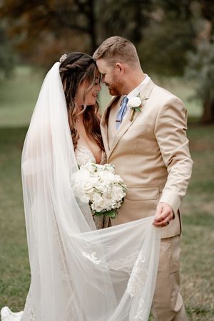 Bride and groom smile tenderly as her lace veil flows in the breeze, groom in tan suit holding white floral bouquet.