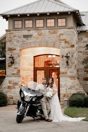 Bride and groom posing with a motorcycle in front of an elegant stone wedding venue at sunset.