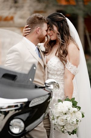 Bride and groom share a romantic moment, bride holding white bouquet in lace gown with veil, groom in beige suit.
