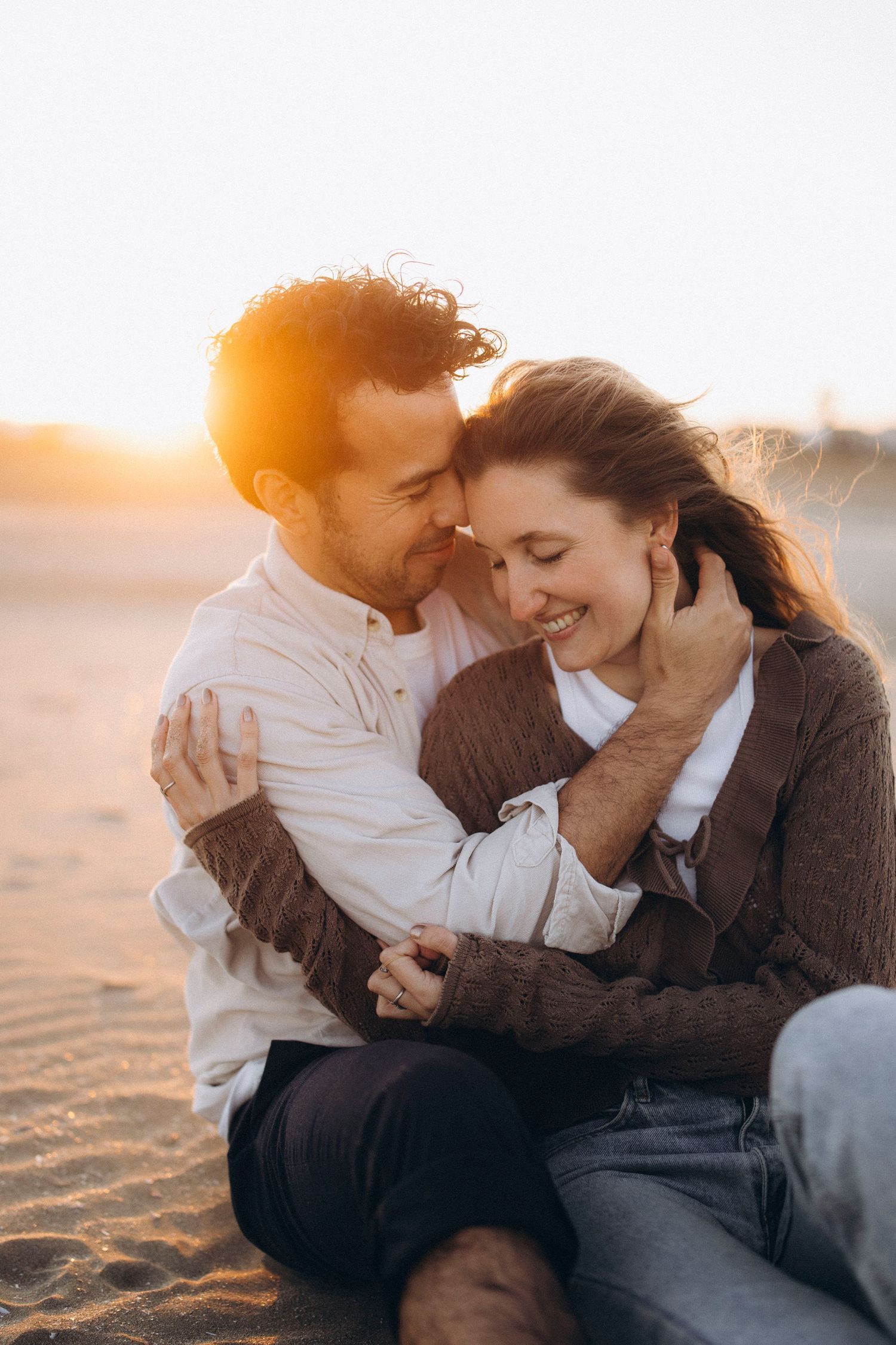 Romantic couple embracing on sandy beach at golden sunset, smiling and holding hands outdoors.