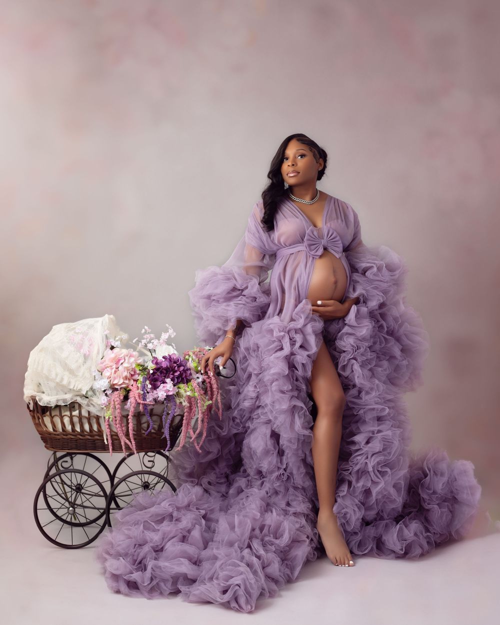 A dreamy maternity photoshoot featuring a flowing purple tulle gown and vintage flower carriage against a soft pink backdrop.