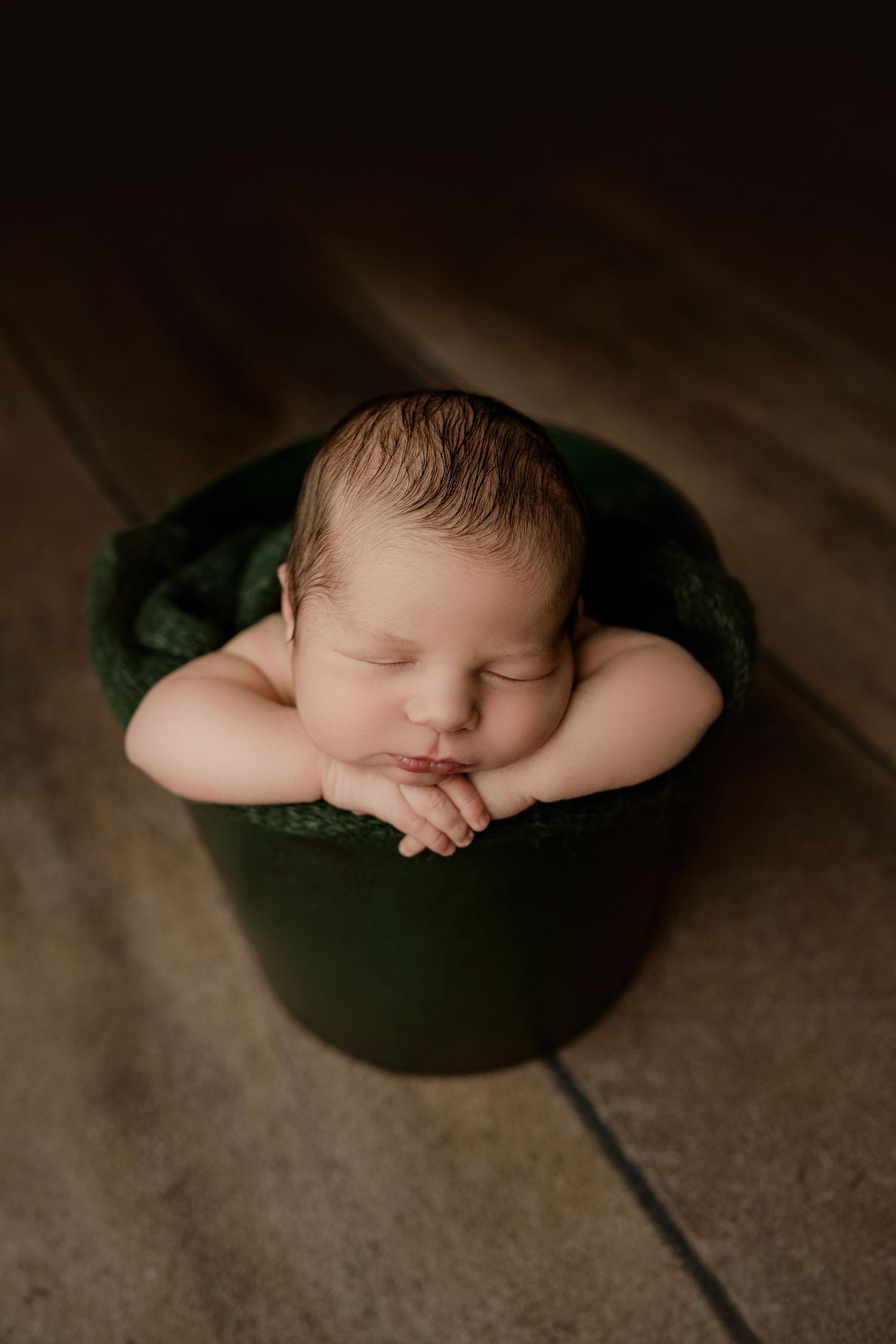 Newborn baby sleeping peacefully in a green bucket with hands tucked under chin on a wooden floor.