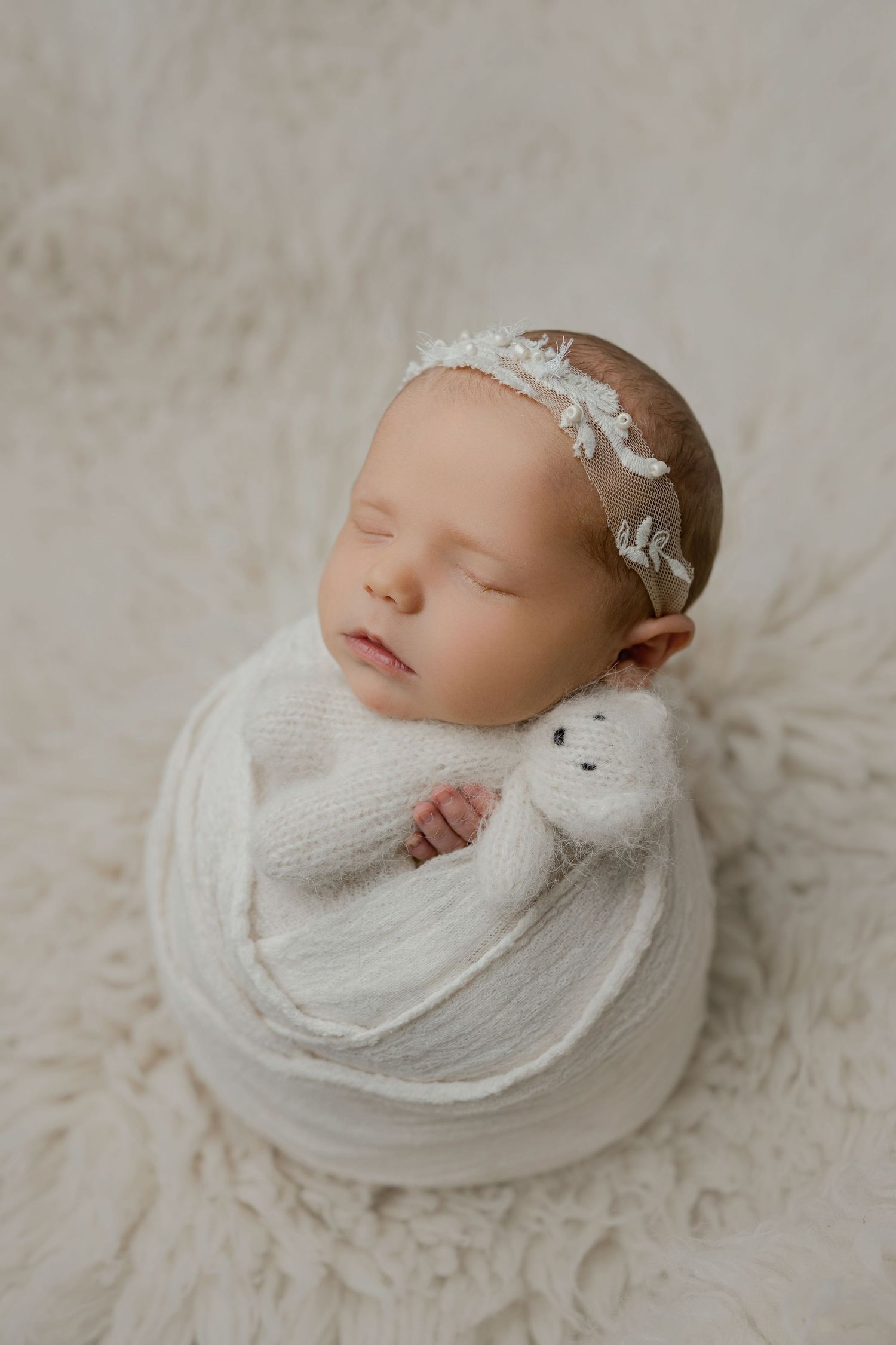 Newborn baby girl swaddled in white wrap holding a stuffed animal, wearing a floral headband, sleeping peacefully.