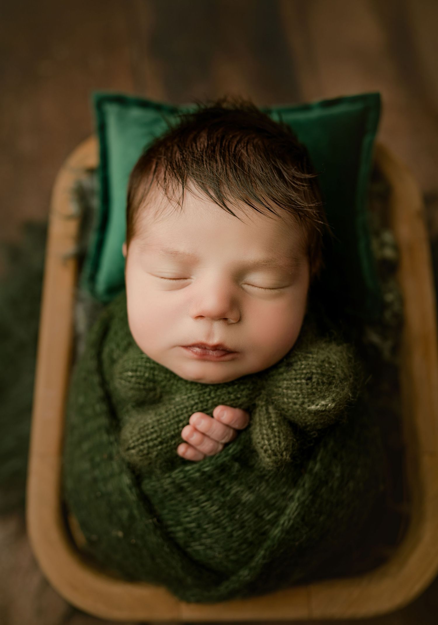 Sleeping newborn baby wrapped in green knit blanket resting in wooden bowl with green velvet pillow.