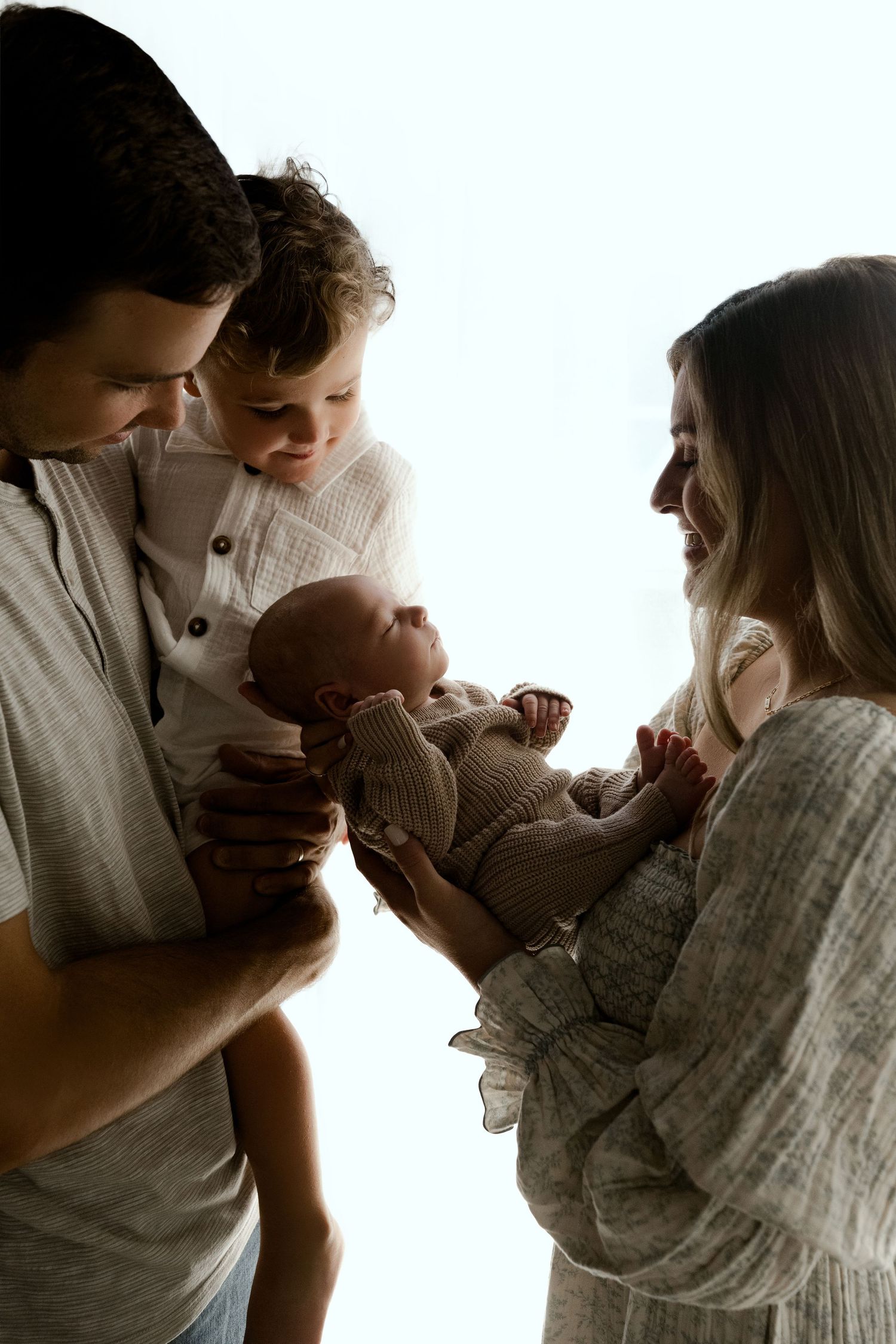 A family of four admires their newborn baby, with parents and toddler gathered closely together in a bright, soft-lit studio.