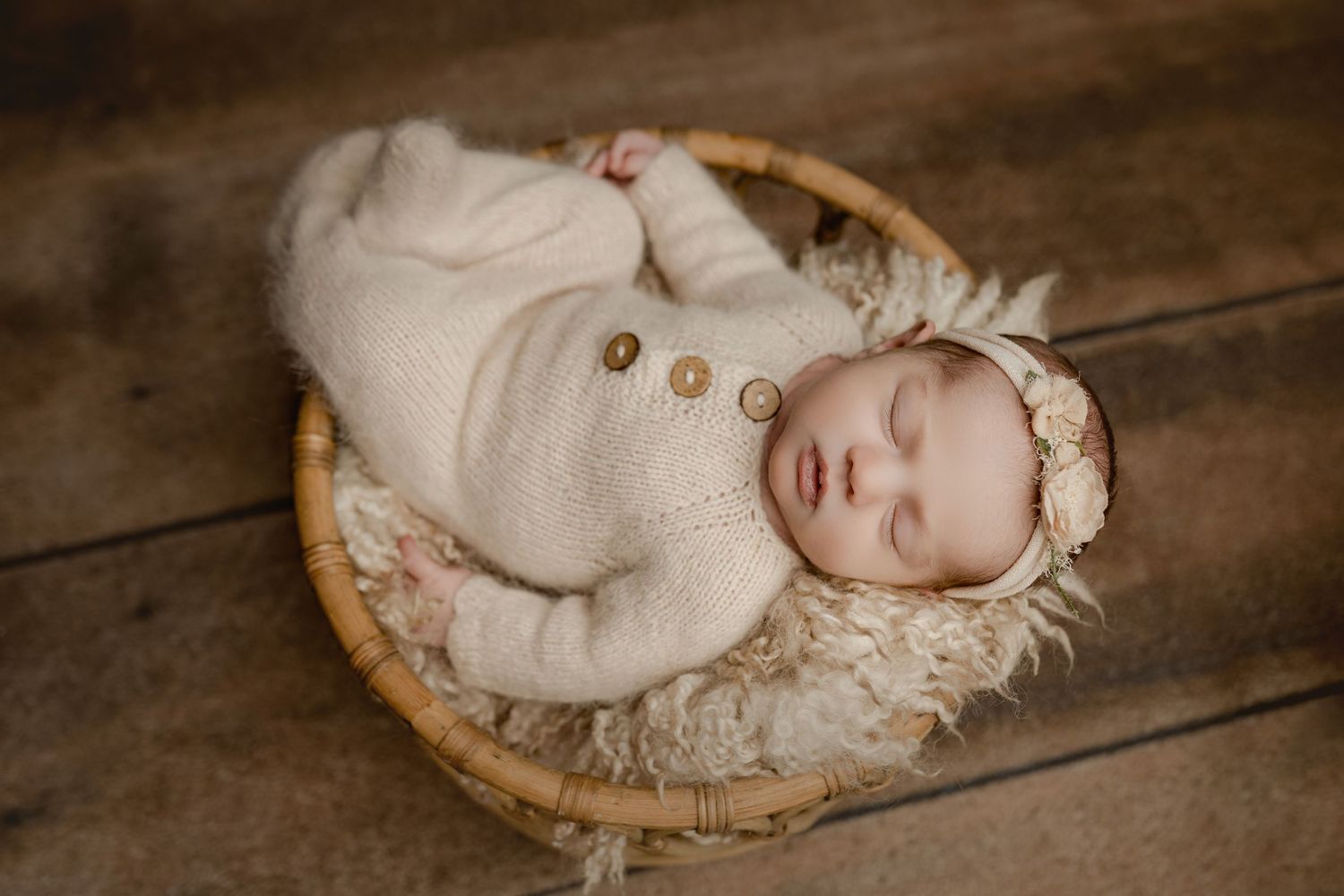 Newborn baby sleeping peacefully in a wicker basket, wearing a cream knit outfit and floral bonnet on a wooden floor.