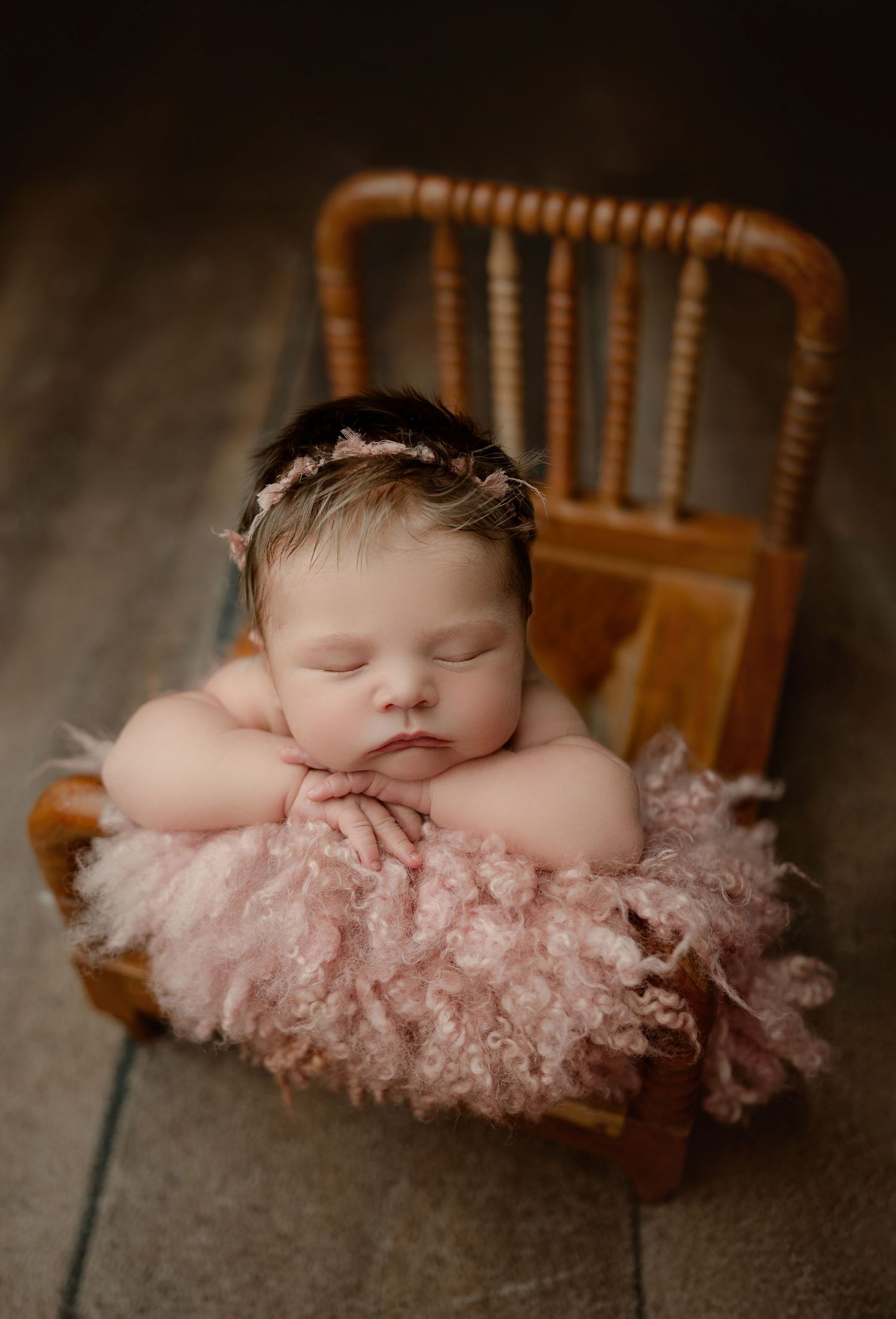 Sleeping newborn baby girl in tiny wooden bed with pink fluffy blanket and floral headband.