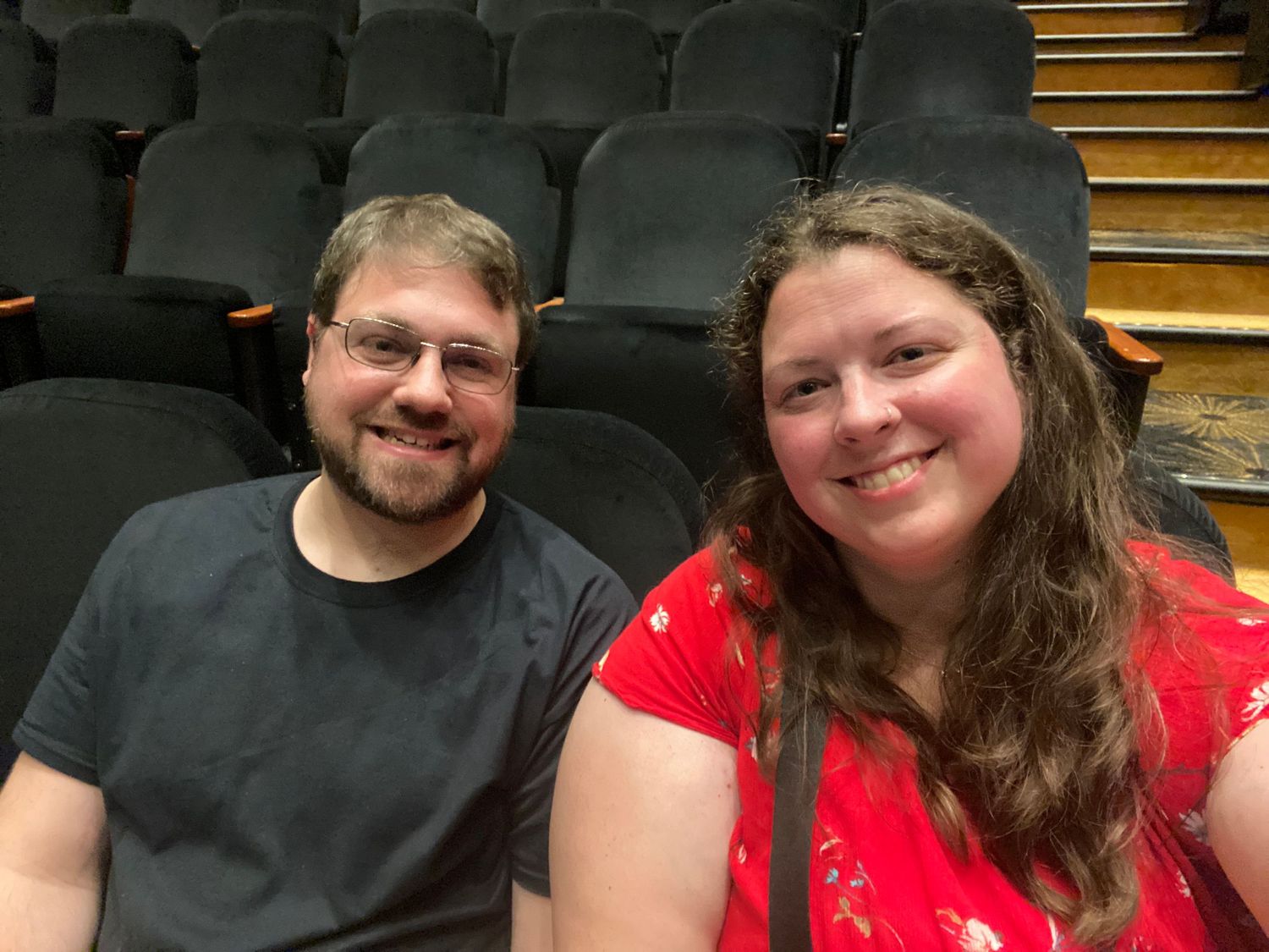 A man and woman smiling together in a theater with rows of empty seats visible in the background.