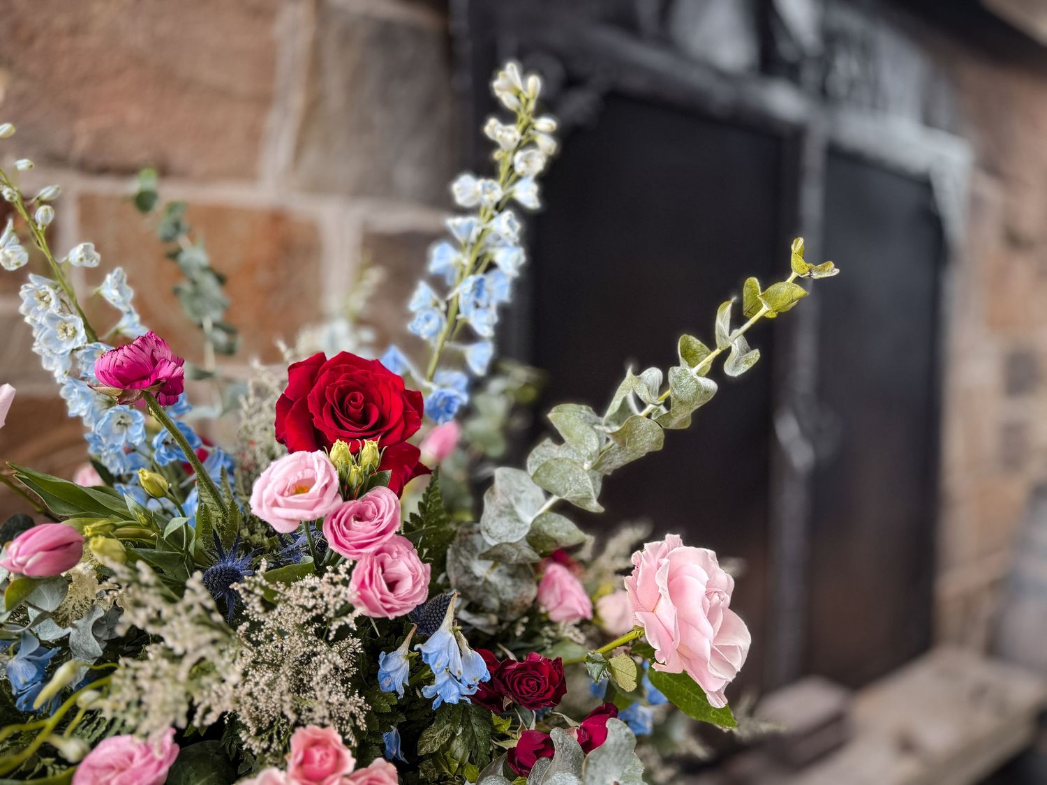 Elegant floral arrangement featuring red, pink roses and blue delphiniums against a rustic brick fireplace backdrop.