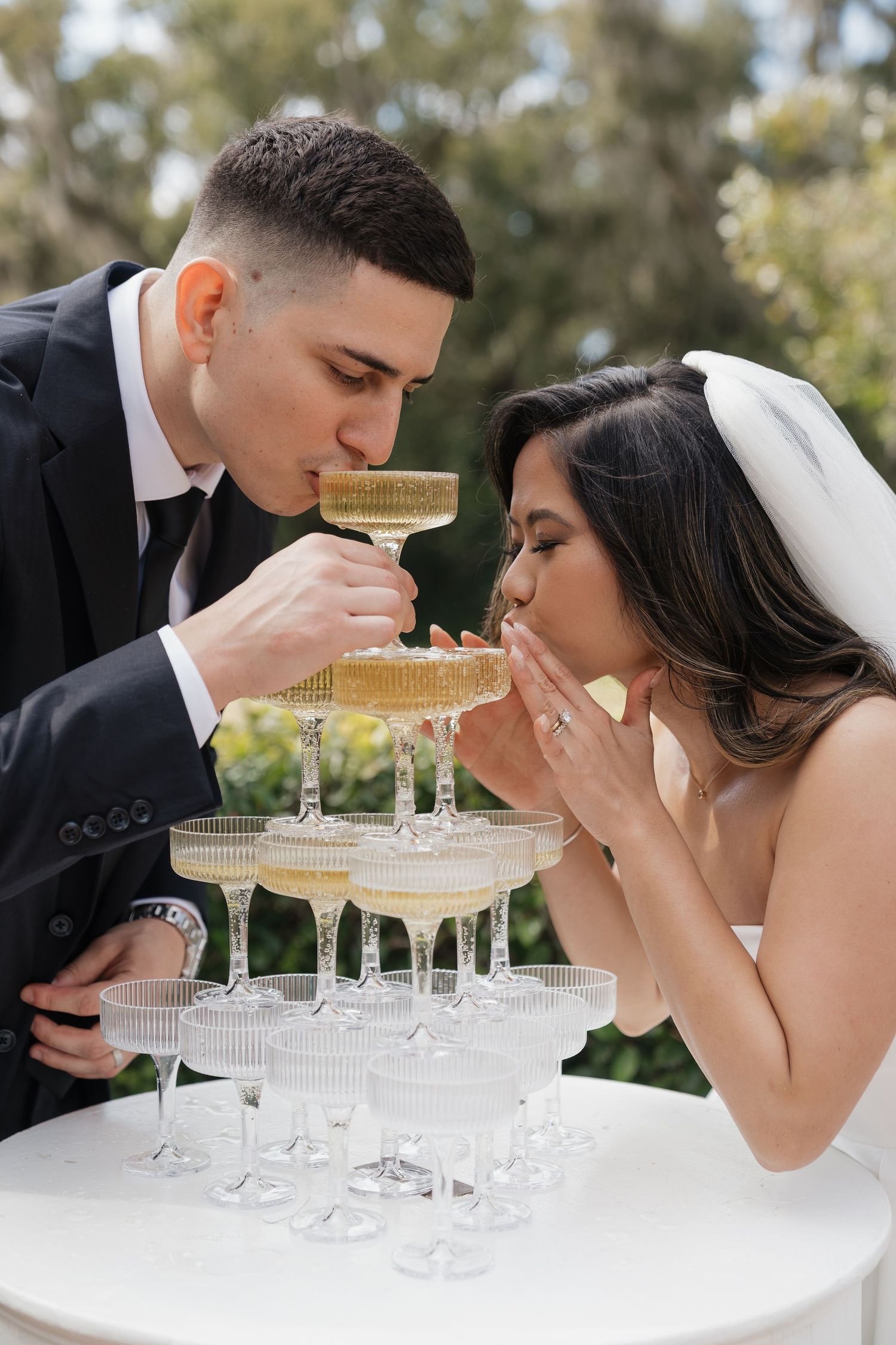 Bride and groom sipping champagne together from a tiered champagne tower at their outdoor wedding reception.