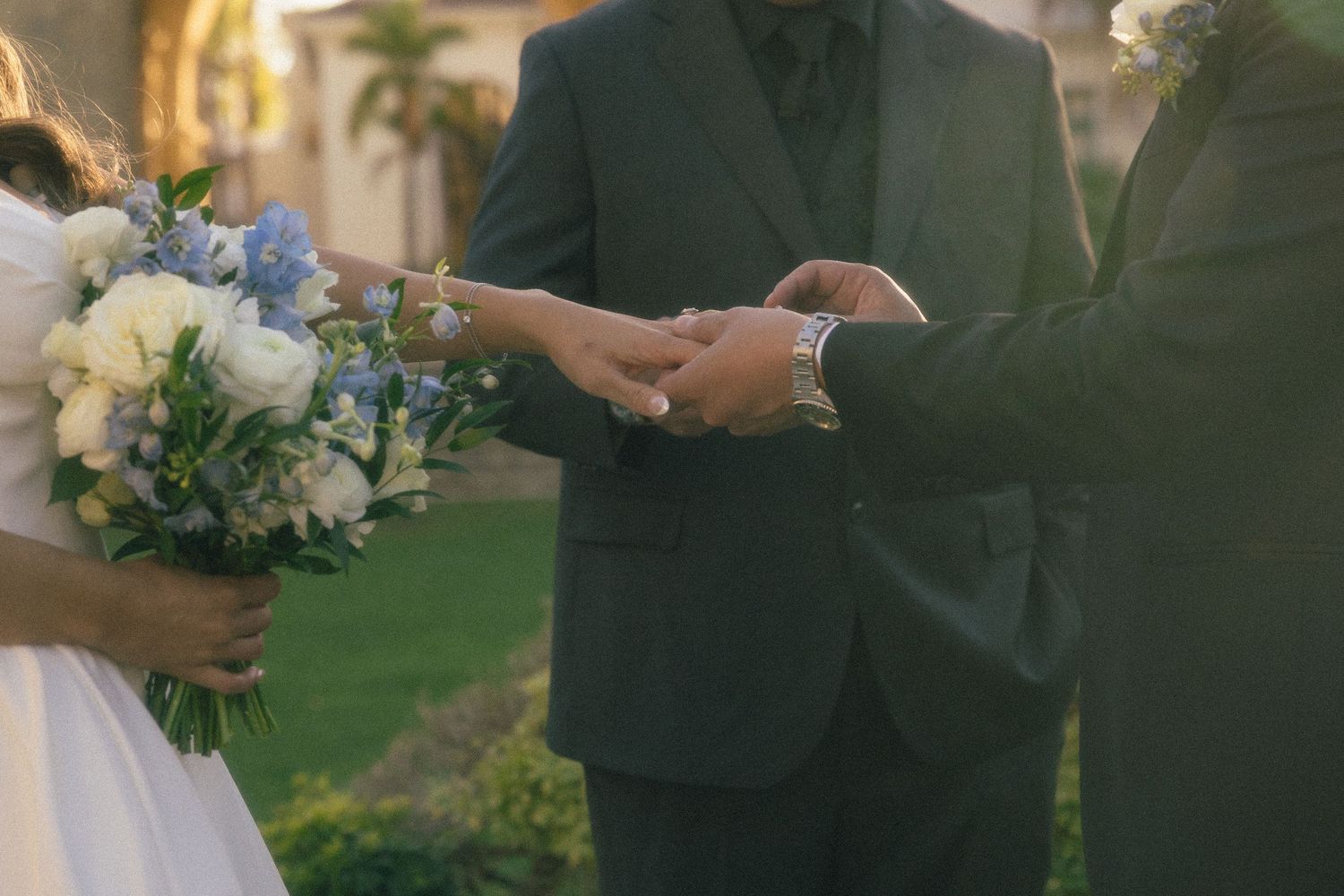 Groom placing wedding ring on bride's finger outdoors, bride holding blue and white floral bouquet.