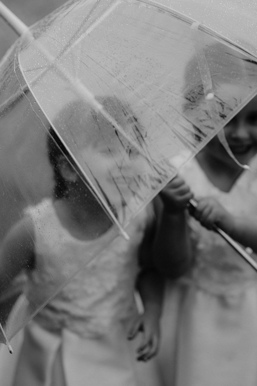 Black and white photo of two girls in white dresses sharing a clear umbrella in the rain.