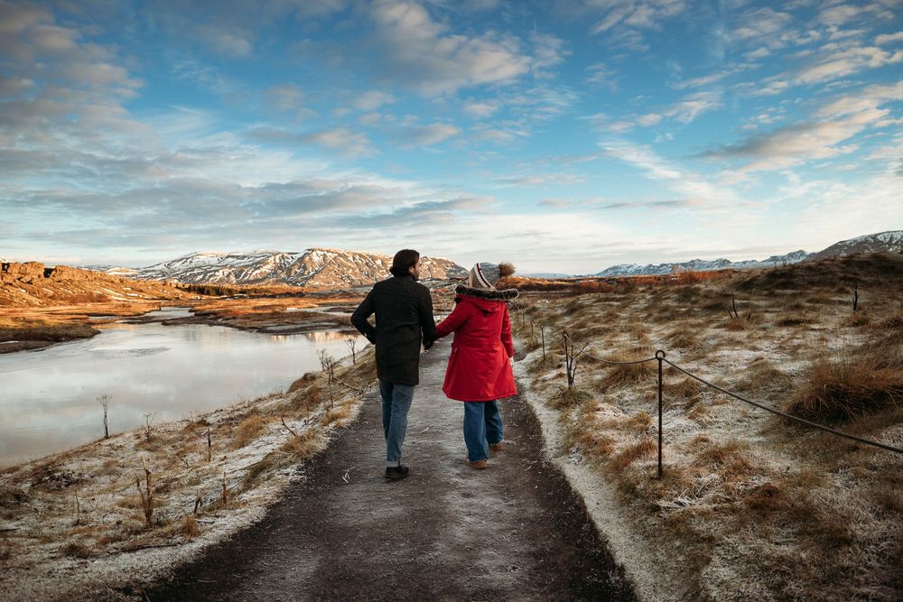 Couple walking hand in hand on a scenic path beside a lake with snow-capped mountains under a dramatic winter sky.
