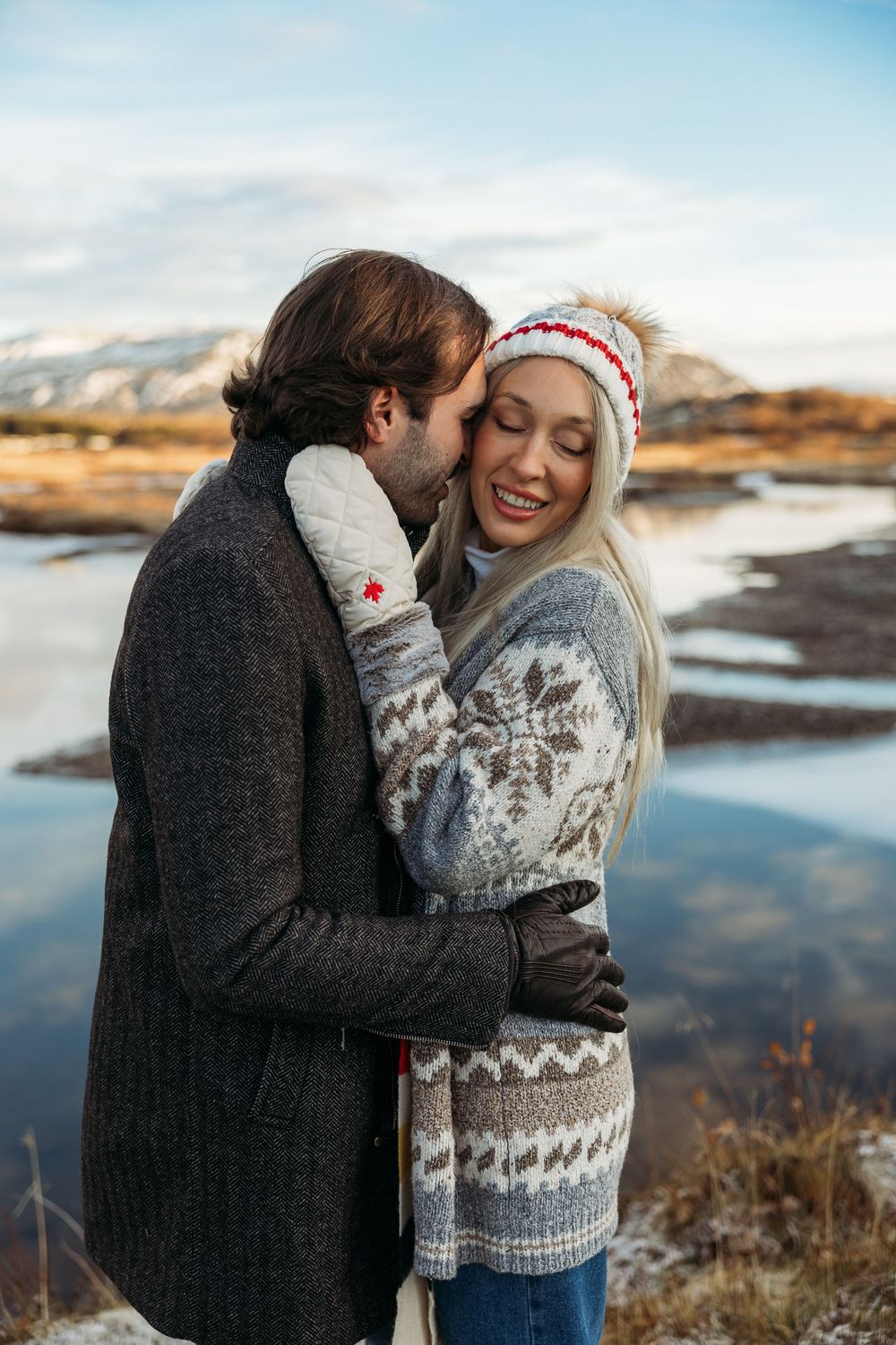 A couple embraces tenderly outdoors in winter, the woman smiling in a Nordic sweater and pom-pom hat near a frozen lake.