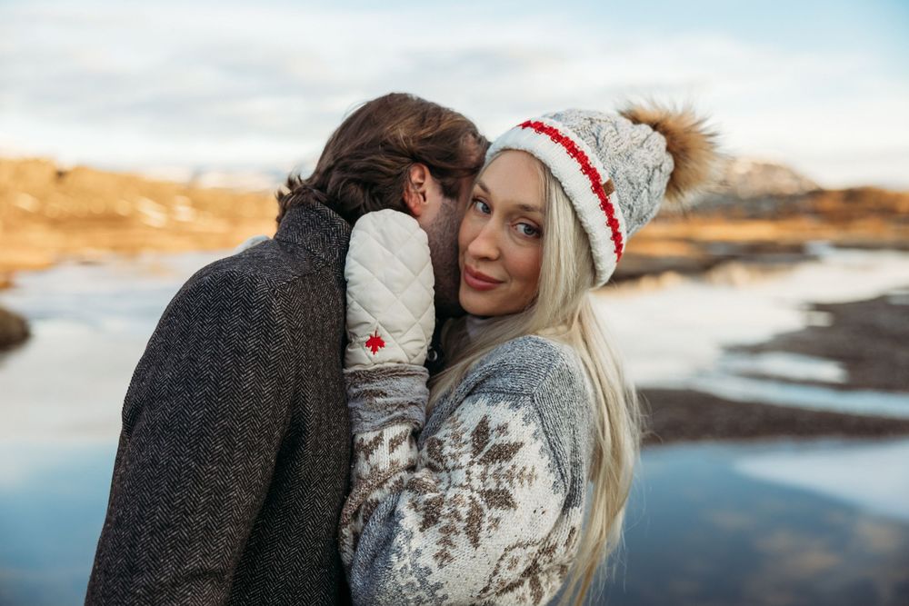 Couple in winter sweaters embracing outdoors near a reflective lake, woman wearing a white and red pom-pom hat.