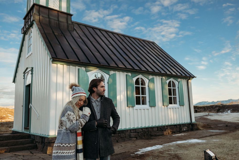 Couple standing together outside a white and teal wooden church in Iceland under a blue sky.