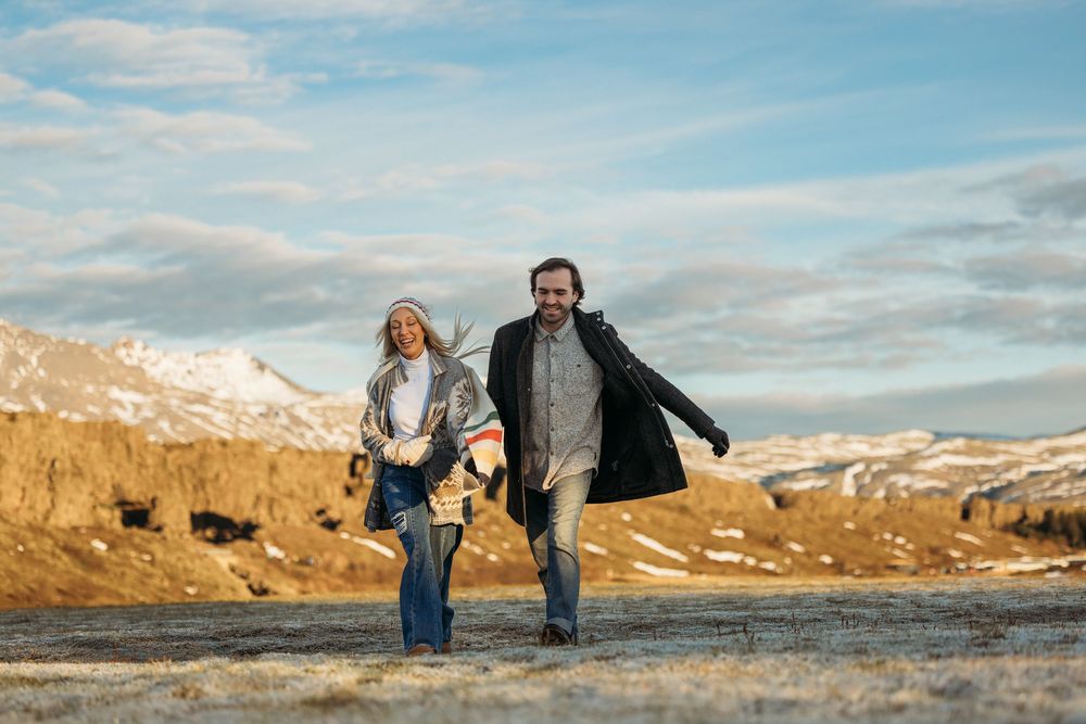 Two people running outdoors in a snowy mountain landscape under a cloudy sky, wearing winter clothing.