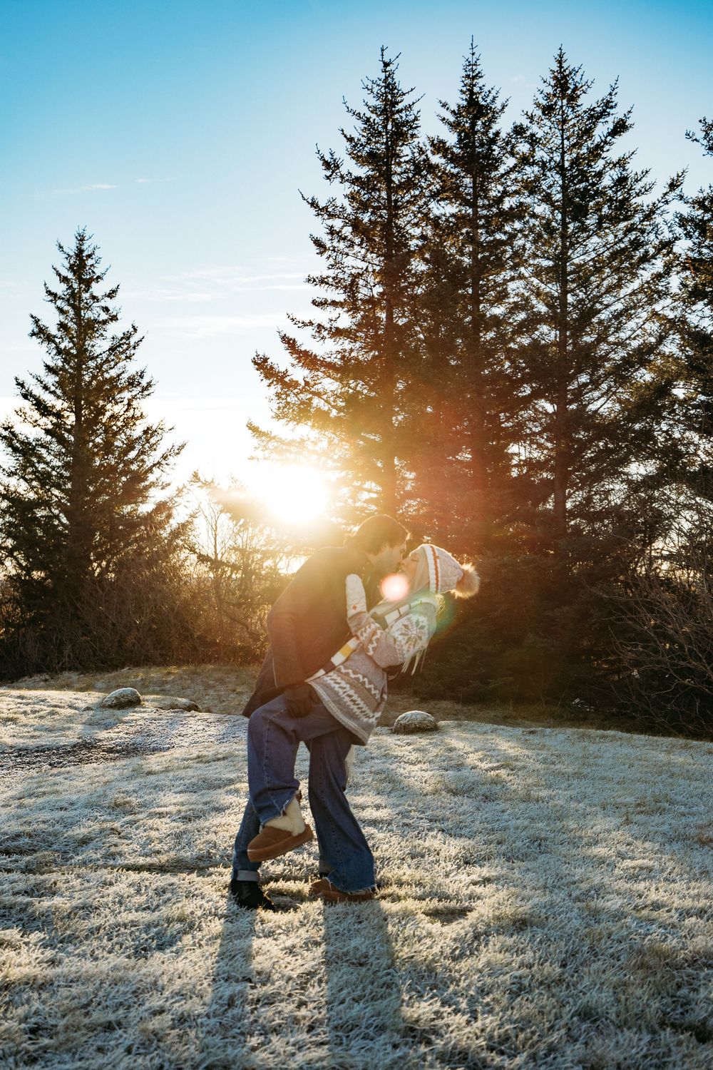 Couple embracing at sunset in a frost-covered field with tall evergreen trees and golden sun flare behind them.