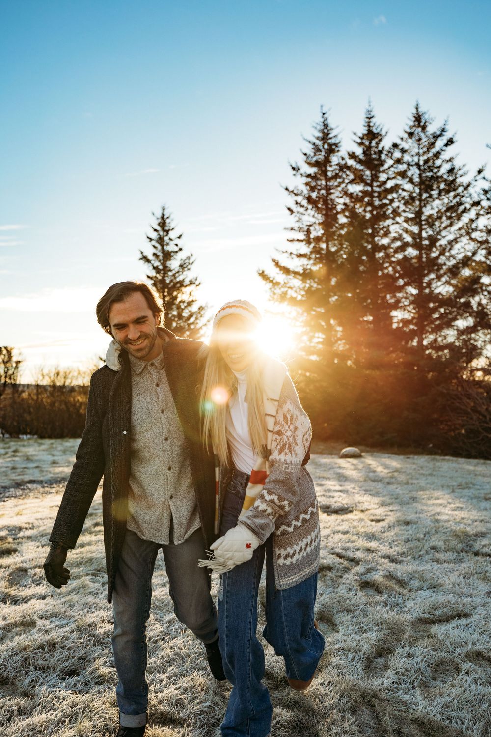 Couple walking together outdoors in winter, smiling at sunset with frost-covered ground and evergreen trees in background.