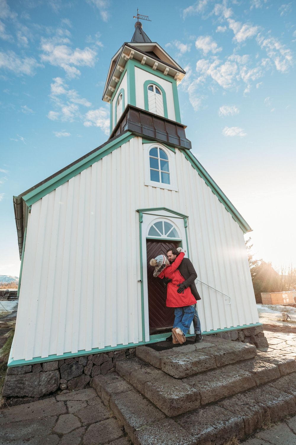 Couple embracing at the entrance of a white wooden church with green trim under a blue sky.