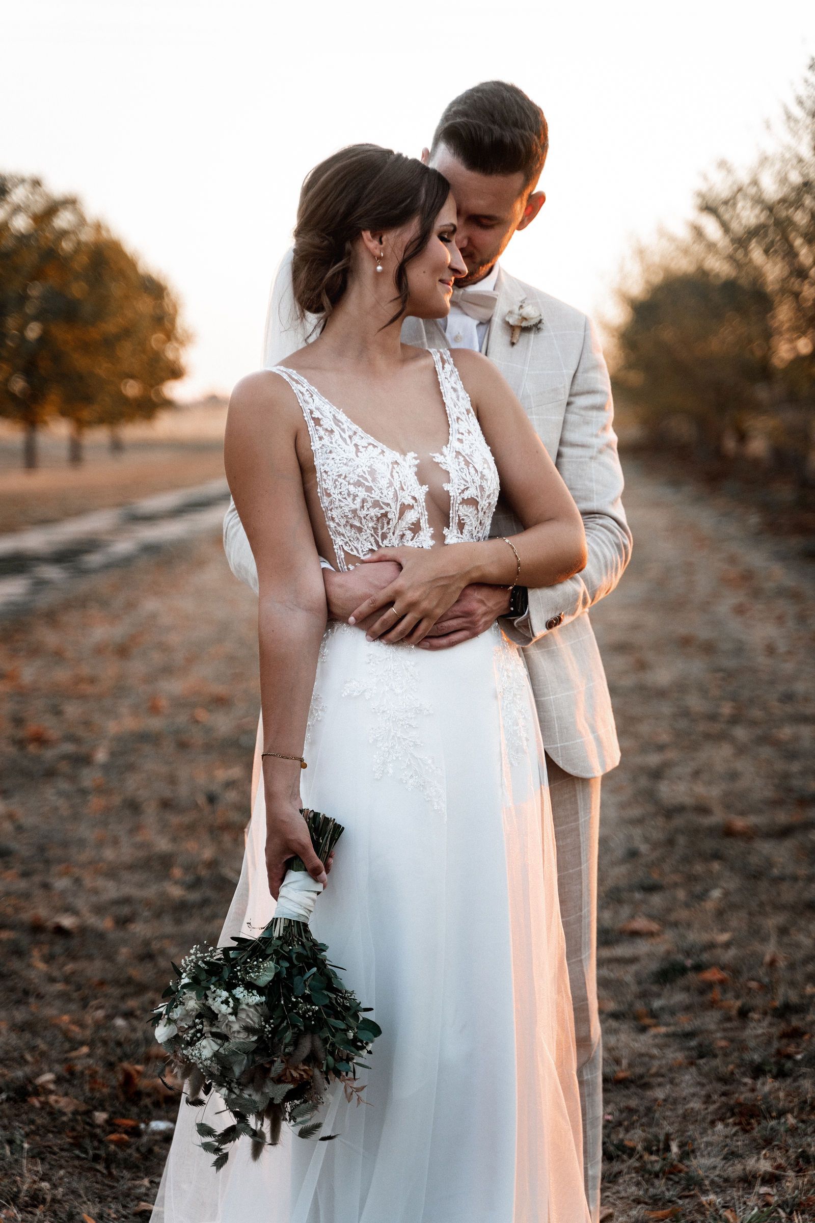 Bride in white gown walks with five bridesmaids in silver satin dresses outdoors on a dirt path.