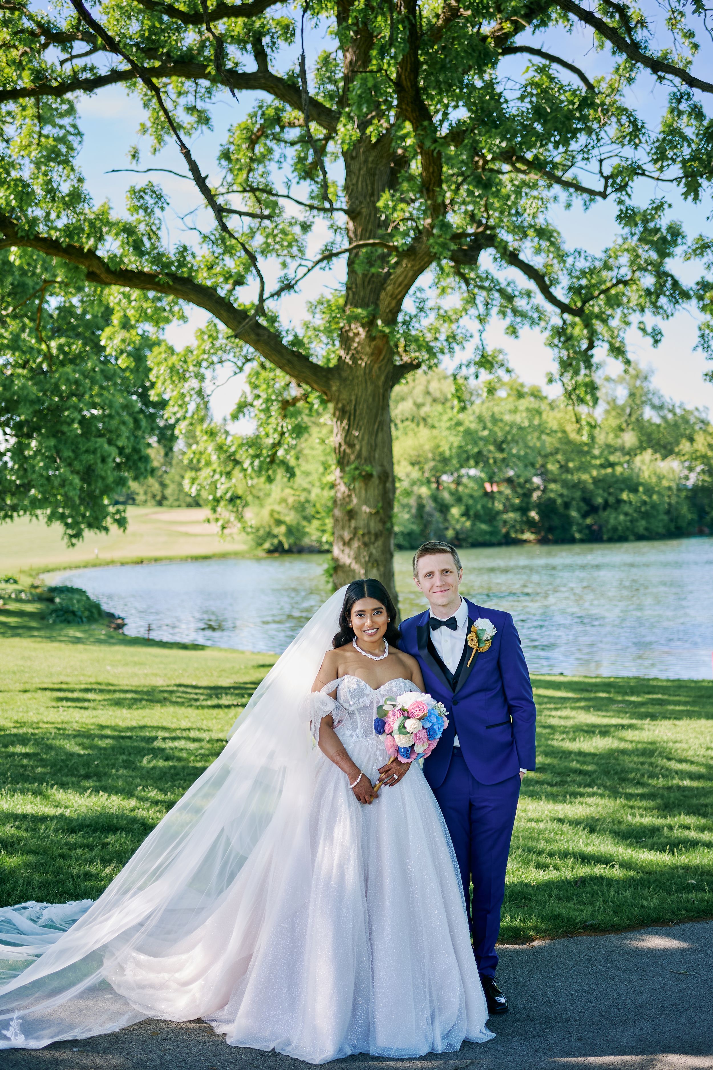 Bride in white ballgown and groom in navy suit pose by a lake under a large tree on their wedding day.