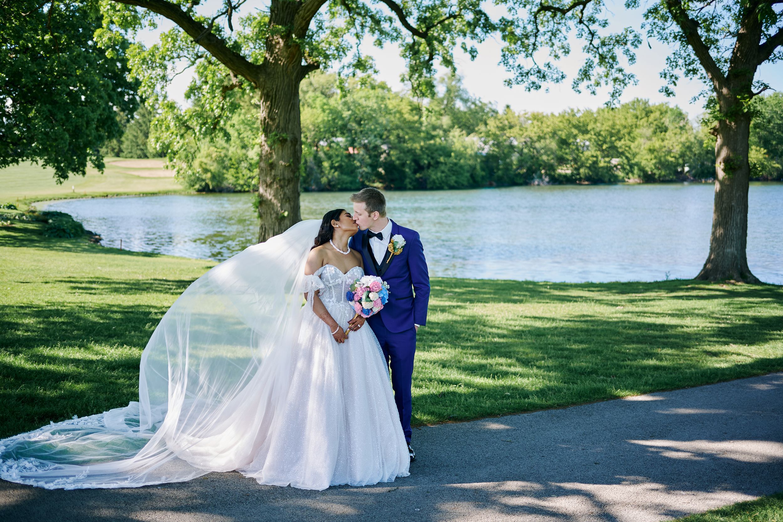 Bride and groom share a kiss by a serene lake, her veil flowing in the breeze, surrounded by lush green trees.