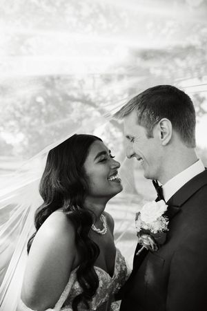 Black and white photo of a smiling bride and groom sharing a joyful moment under a wedding veil.