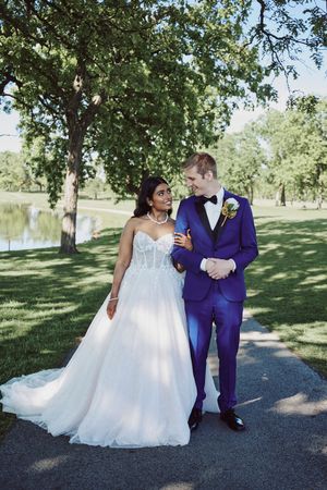 Bride in white gown and groom in blue suit walk together in a lush green park on their wedding day.
