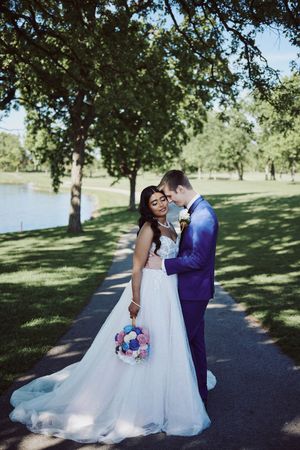 Bride in white gown holding colorful bouquet stands with groom in purple suit in a sunny park near a lake.