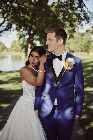 Bride in white gown and groom in blue suit pose together outdoors near a lake surrounded by trees.