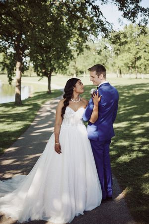 Bride in white gown and groom in blue suit share a romantic moment on a sunlit park path near a pond.