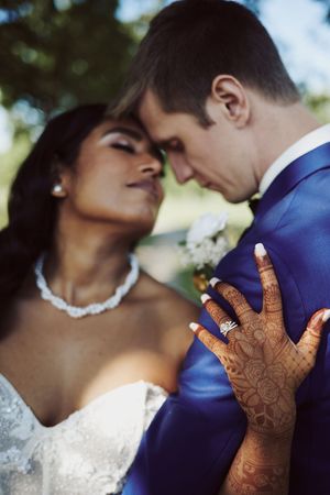 Interracial couple sharing romantic moment, bride with henna hands and diamond ring in white gown, groom in blue suit.