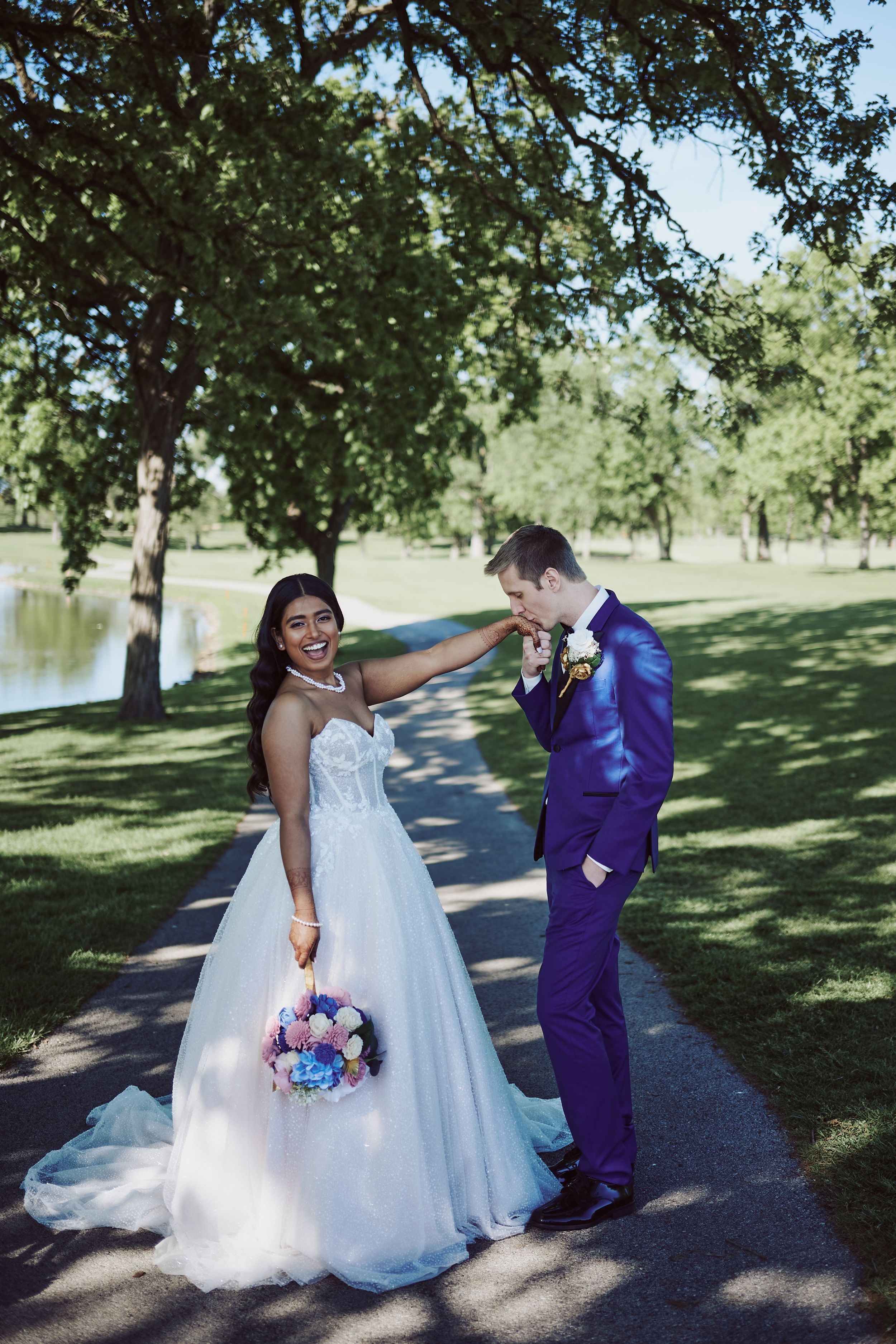 Bride in white gown holds colorful bouquet as groom in purple suit kisses her hand in a lush green park.