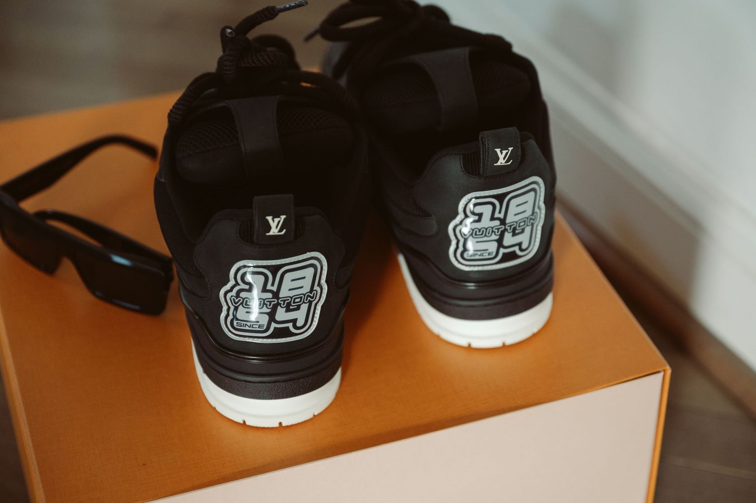 Black Louis Vuitton LV Trainer sneakers displayed on an orange box with sunglasses beside them.