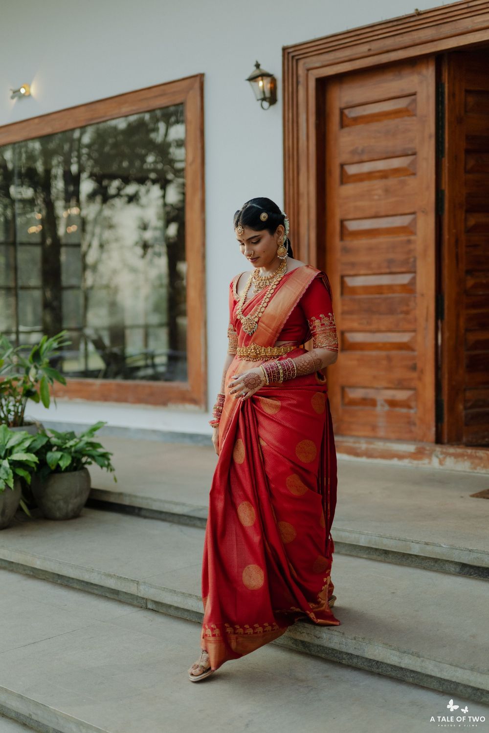 South Indian bride in red Kanjivaram silk saree with gold jewelry poses gracefully outside a white building.
