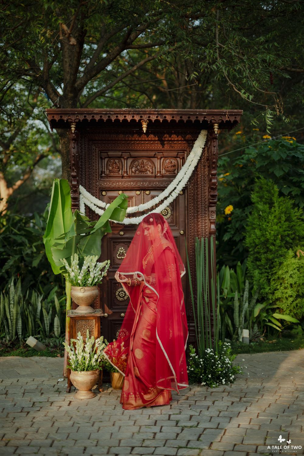 South Indian bride in red saree and veil stands before ornate carved wooden temple door in garden.