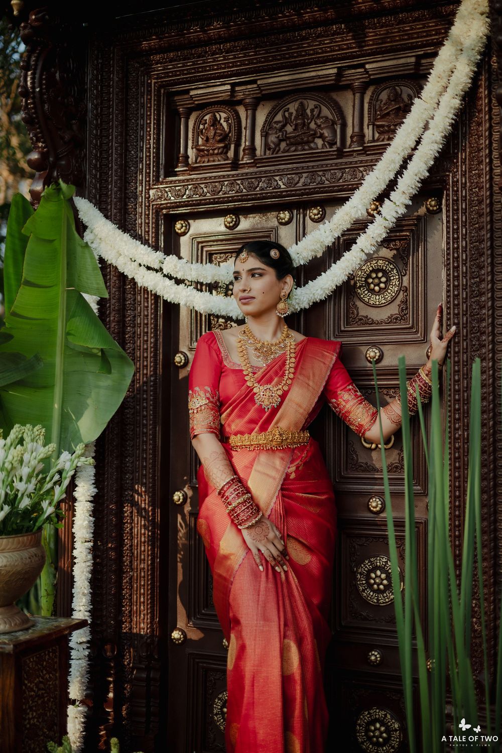 South Indian bride in red Kanjivaram saree poses by intricately carved wooden door with white floral garlands.
