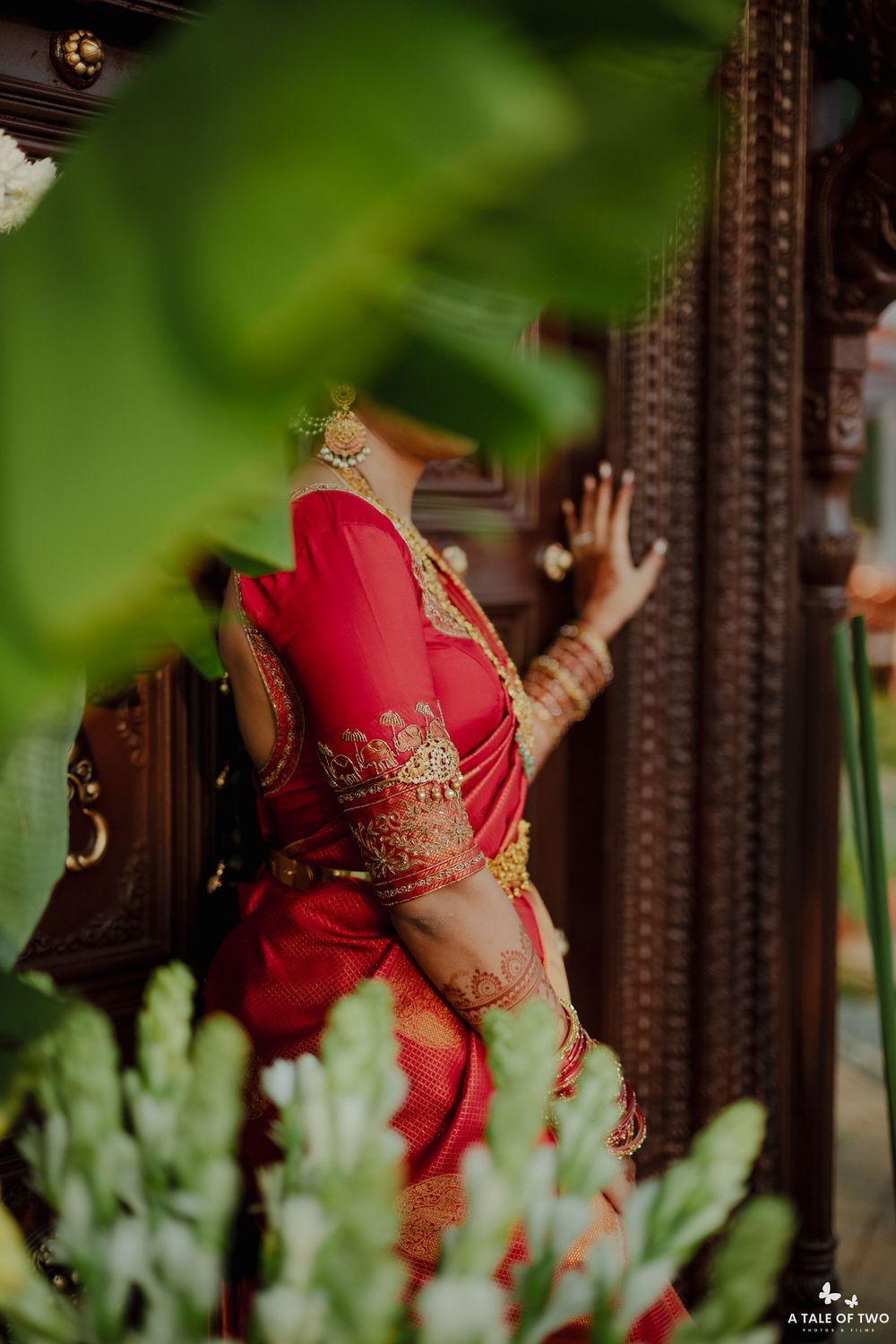 Bride in red silk saree with gold embroidery poses beside carved wooden temple door framed by green leaves.