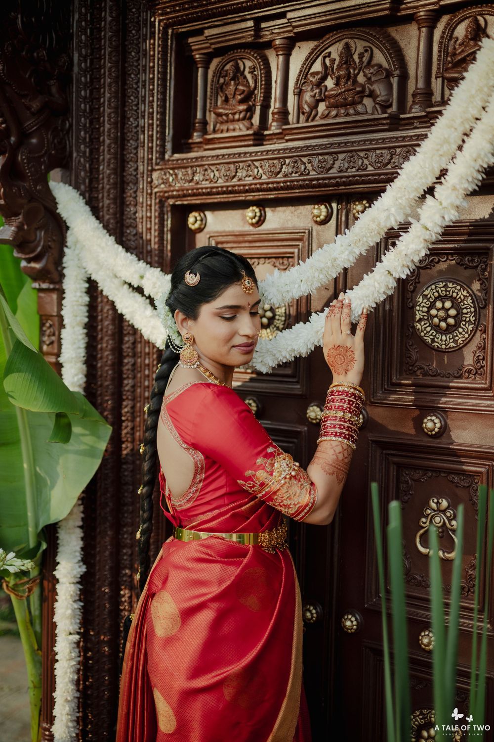 South Indian bride in red Kanjivaram saree touches ornate carved wooden temple door with mehndi hands.