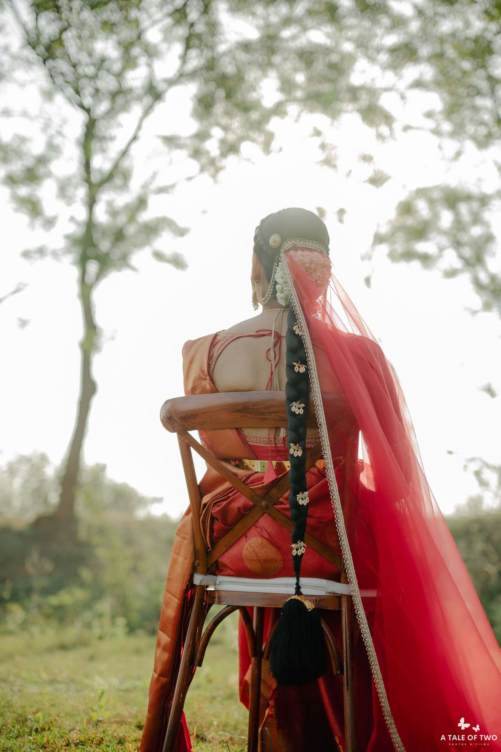 Bride in red saree with floral braid sits on chair outdoors amid misty trees.