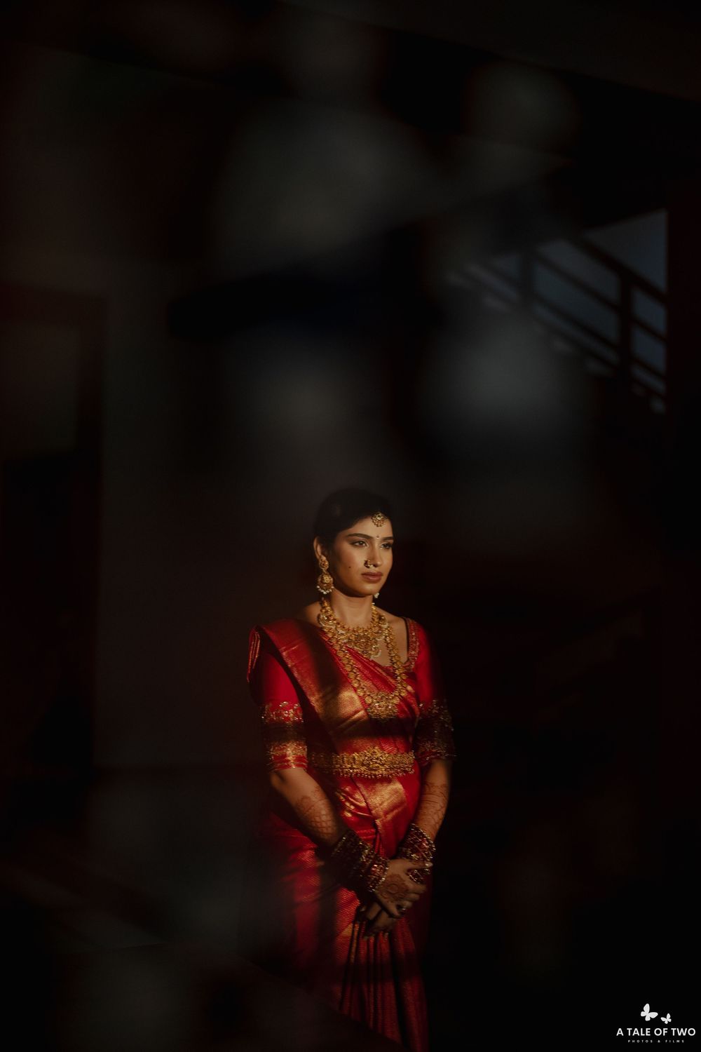 Bride in red silk saree with gold jewelry stands in dramatic light during South Indian wedding ceremony.