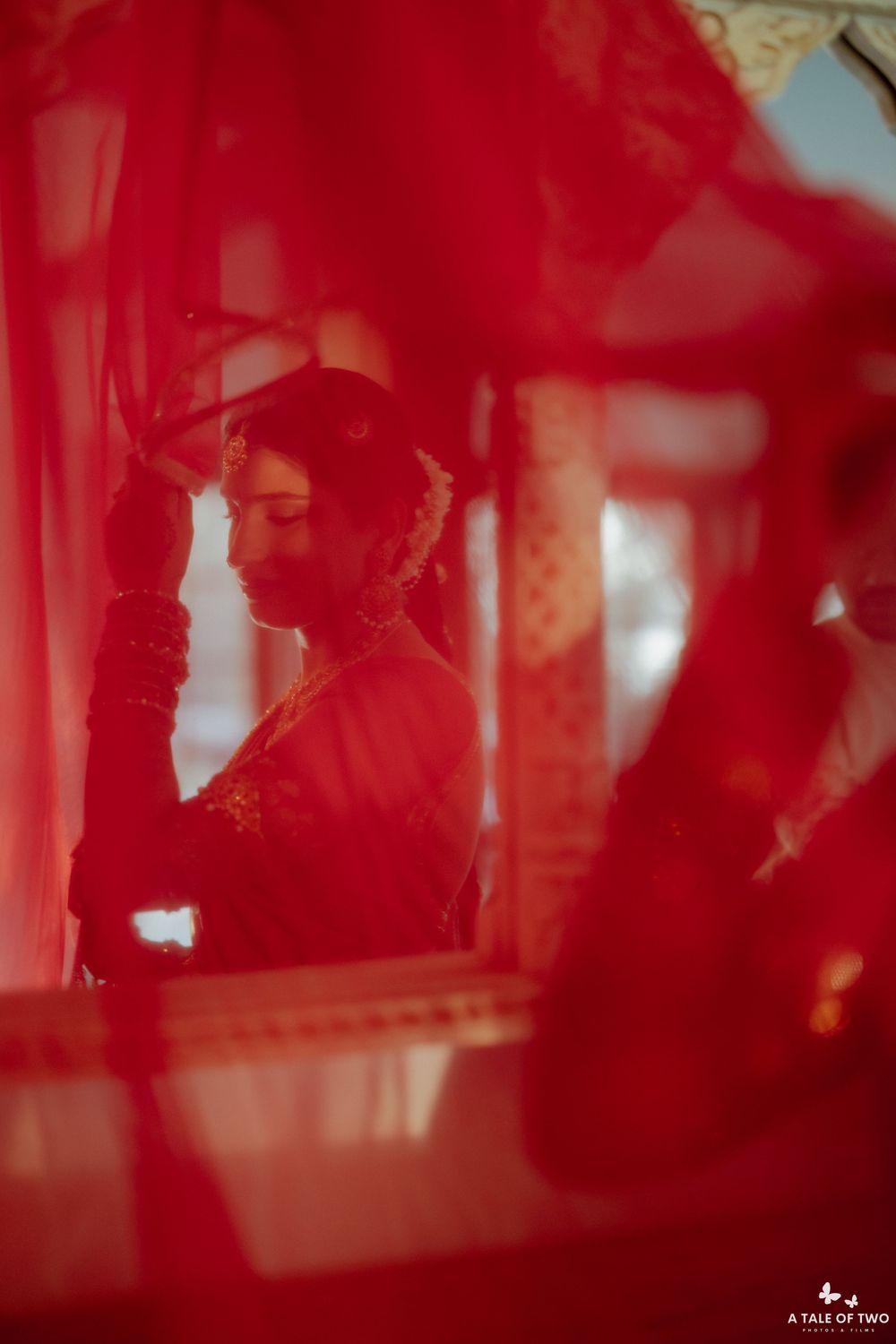 Bride in red saree seen through sheer red drapes during Indian wedding preparation.