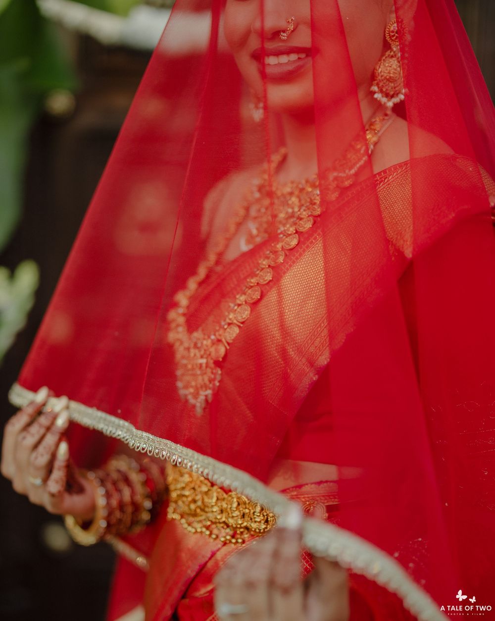 Close-up of Indian bride in red saree with gold necklace and bangles holding white garland under red veil.